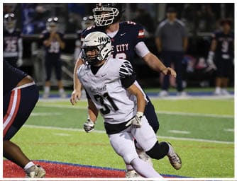 A group of people in football uniforms are actively playing on a field with one person in white running while others in dark uniforms are nearby. Descripci n generada con IA