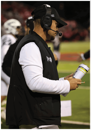 Person wearing a headset and a black vest over a white long-sleeve shirt is looking at a clipboard, likely coaching or managing a sports event. Descripci n generada con IA