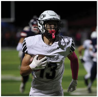 A person wearing a white football uniform and helmet is running while holding a football on a field during a nighttime game. Descripci n generada con IA