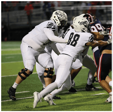 Two people in white football uniforms with helmets labeled “HAWKS" are actively engaged in blocking opponents on a football field during a game. Descripci n generada con IA