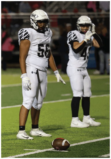 Two people in white football uniforms and helmets stand on a green field during a game, with one person holding their face and the other standing with hands by their side. Descripci n generada con IA