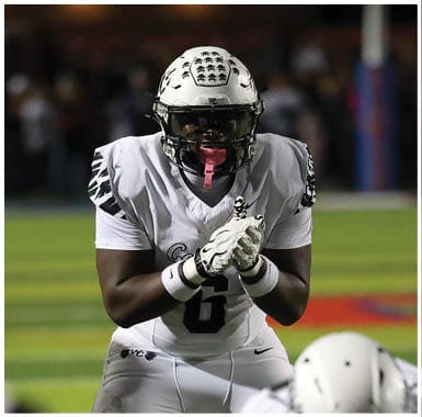 A person in a white football uniform with the number 6 is positioned behind the line of scrimmage, preparing to receive the snap during a nighttime football game. Descripci n generada con IA