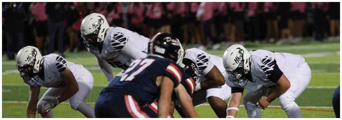 People in football uniforms are lined up on a field preparing for a play with one person holding the football on the ground. Descripci n generada con IA