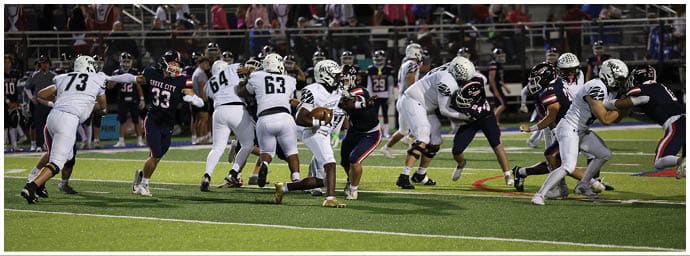 People wearing football uniforms are actively engaged in a play on a green field with a crowd in the background. Descripci n generada con IA