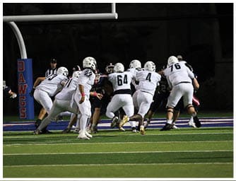 A group of football players in white uniforms are lined up on the field facing opponents in dark uniforms during a nighttime game. Descripci n generada con IA