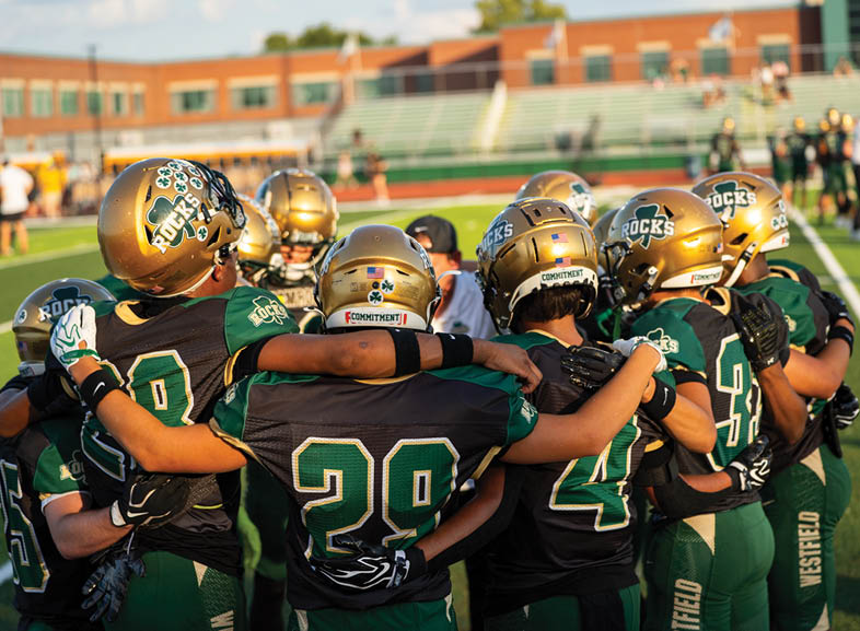 Four people wearing black football uniforms with “VISTA" on the front are standing together in a stadium with a crowd in the background, two of them holding helmets raised above their heads. Description generated by AI