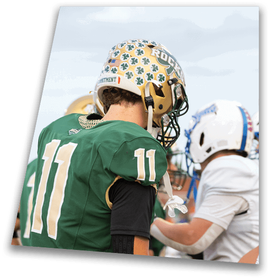 A person in a black football uniform and helmet is crouched in a ready position on a green field with another player in white uniform visible in the background. Description generated by AI