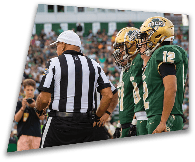 Four people in dark football uniforms and helmets stand in a row on a field, appearing focused and ready for action. Description generated by AI
