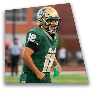 A person in football gear and helmet is crouched on a field with other players lined up in the background during a game. Description generated by AI