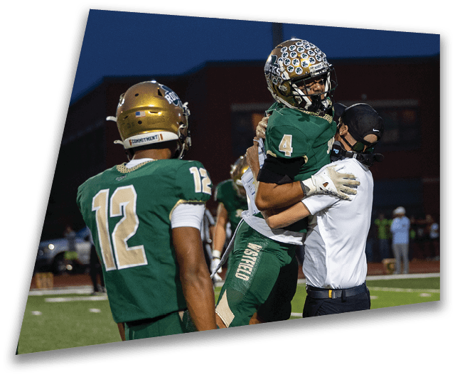 Four people in dark football uniforms and helmets stand in a row on a field, appearing focused and ready for action. Description generated by AI