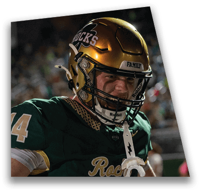 A person in football gear and helmet is crouched on a field with other players lined up in the background during a game. Description generated by AI
