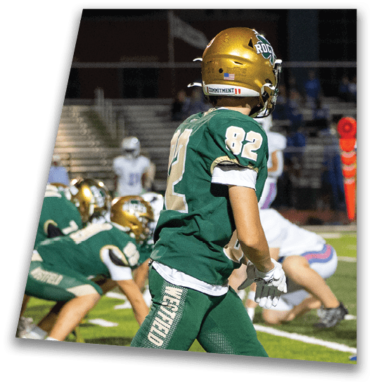 A person in a black football uniform and helmet is crouched in a ready position on a green field with another player in white uniform visible in the background. Description generated by AI