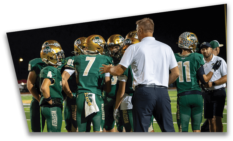 People wearing black American Canyon football uniforms and helmets are standing on a football field at night. Description generated by AI
