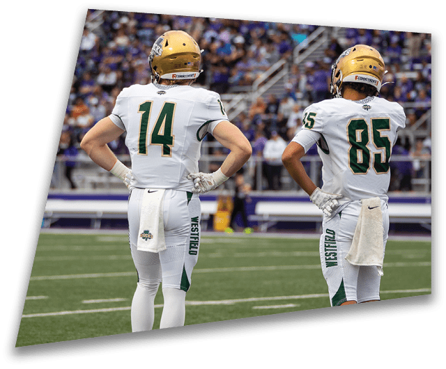 Four people in dark football uniforms and helmets stand in a row on a field, appearing focused and ready for action. Description generated by AI