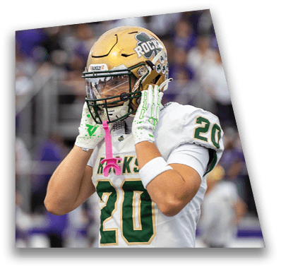 A person in football gear and helmet is crouched on a field with other players lined up in the background during a game. Description generated by AI