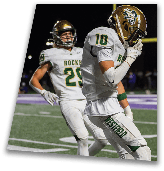 A person in a black football uniform and helmet is crouched in a ready position on a green field with another player in white uniform visible in the background. Description generated by AI