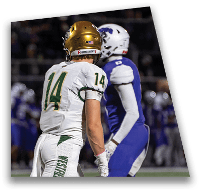 A person in football gear and helmet is crouched on a field with other players lined up in the background during a game. Description generated by AI