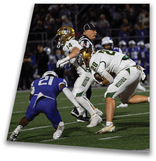 A person in a black football uniform and helmet is crouched in a ready position on a green field with another player in white uniform visible in the background. Description generated by AI