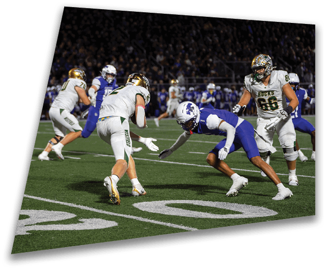 Four people in dark football uniforms and helmets stand in a row on a field, appearing focused and ready for action. Description generated by AI