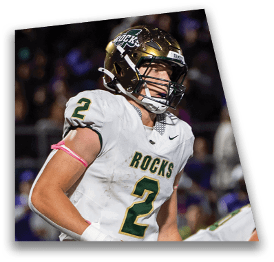 A person in football gear and helmet is crouched on a field with other players lined up in the background during a game. Description generated by AI