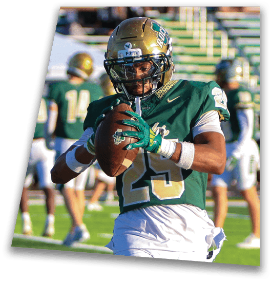 A person in a black football uniform and helmet is crouched in a ready position on a green field with another player in white uniform visible in the background. Description generated by AI