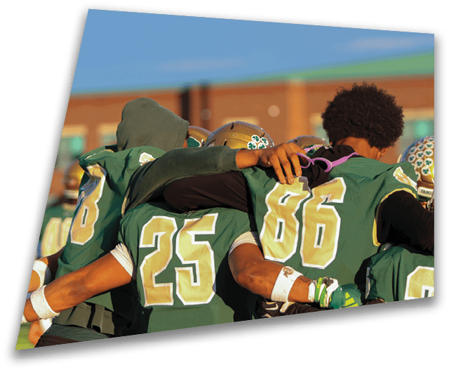 Four people in dark football uniforms and helmets stand in a row on a field, appearing focused and ready for action. Description generated by AI