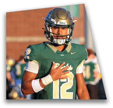 A person in football gear and helmet is crouched on a field with other players lined up in the background during a game. Description generated by AI