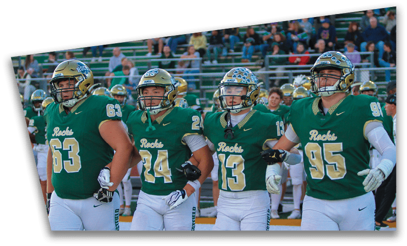 People wearing black American Canyon football uniforms and helmets are standing on a football field at night. Description generated by AI