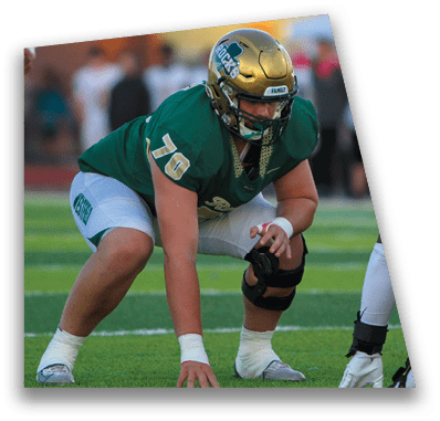 A person in football gear and helmet is crouched on a field with other players lined up in the background during a game. Description generated by AI