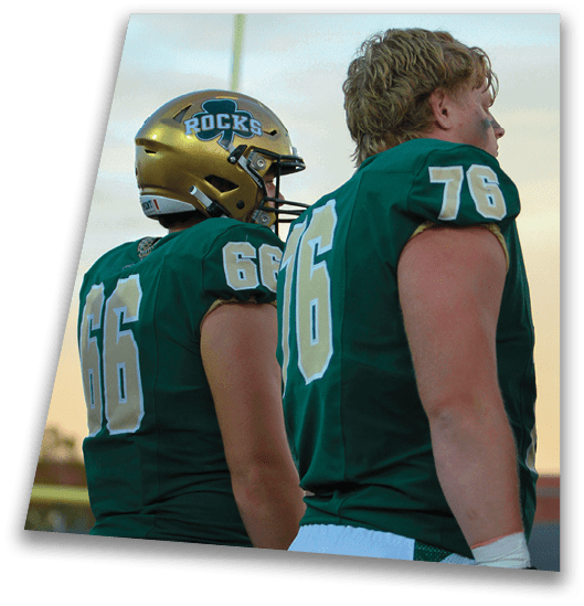 A person in a black football uniform and helmet is crouched in a ready position on a green field with another player in white uniform visible in the background. Description generated by AI