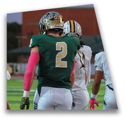 A person in football gear and helmet is crouched on a field with other players lined up in the background during a game. Description generated by AI