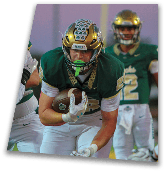 A person in a black football uniform and helmet is crouched in a ready position on a green field with another player in white uniform visible in the background. Description generated by AI