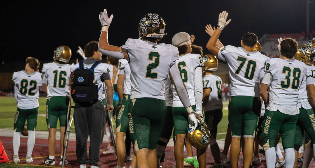 Four people wearing black football uniforms with “VISTA" on the front are standing together in a stadium with a crowd in the background, two of them holding helmets raised above their heads. Description generated by AI