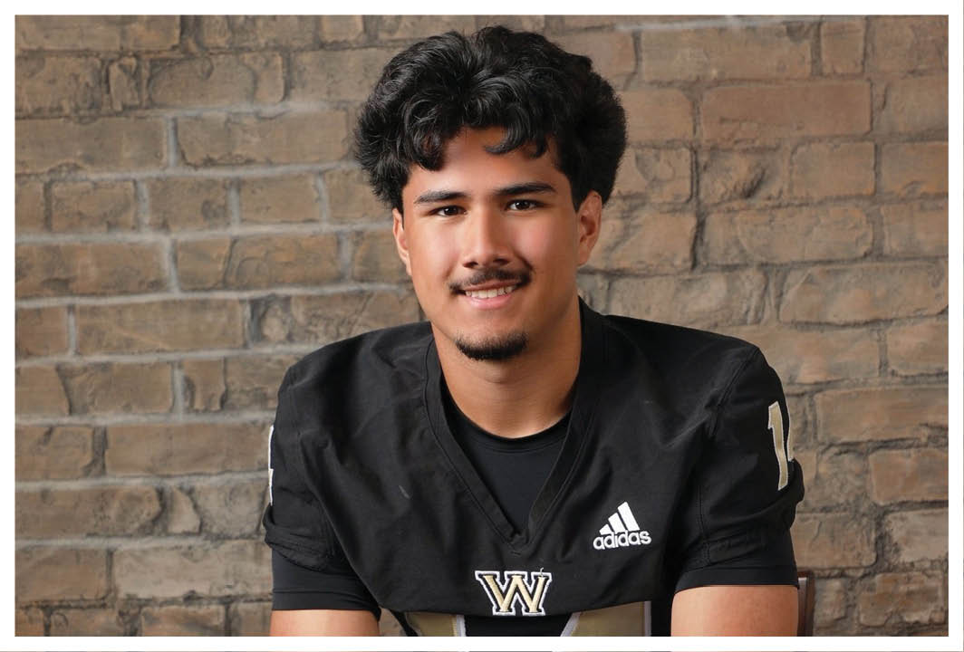 A person with curly hair wearing a black sports jersey with a “W" logo is smiling in front of a brick wall. Description generated by AI