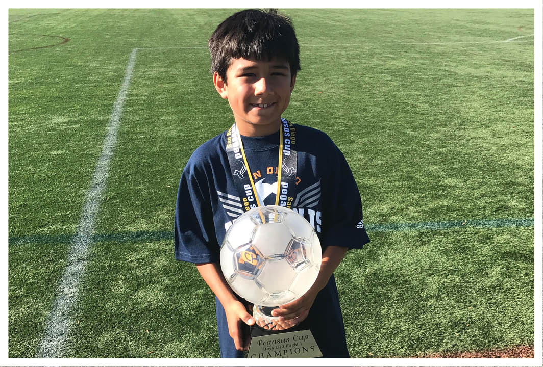 A child wearing a dark sports shirt stands on a grassy field holding a soccer ball and wearing a medal around their neck. Description generated by AI