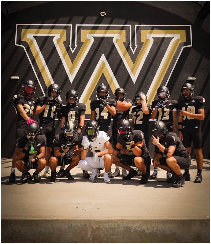 A group of people wearing black football uniforms and helmets pose together in front of a large black and gold wall with a stylized “W" logo. Description generated by AI
