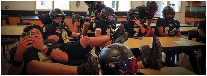 A group of people wearing football helmets and uniforms are sitting and lounging in a classroom with desks and windows. Description generated by AI