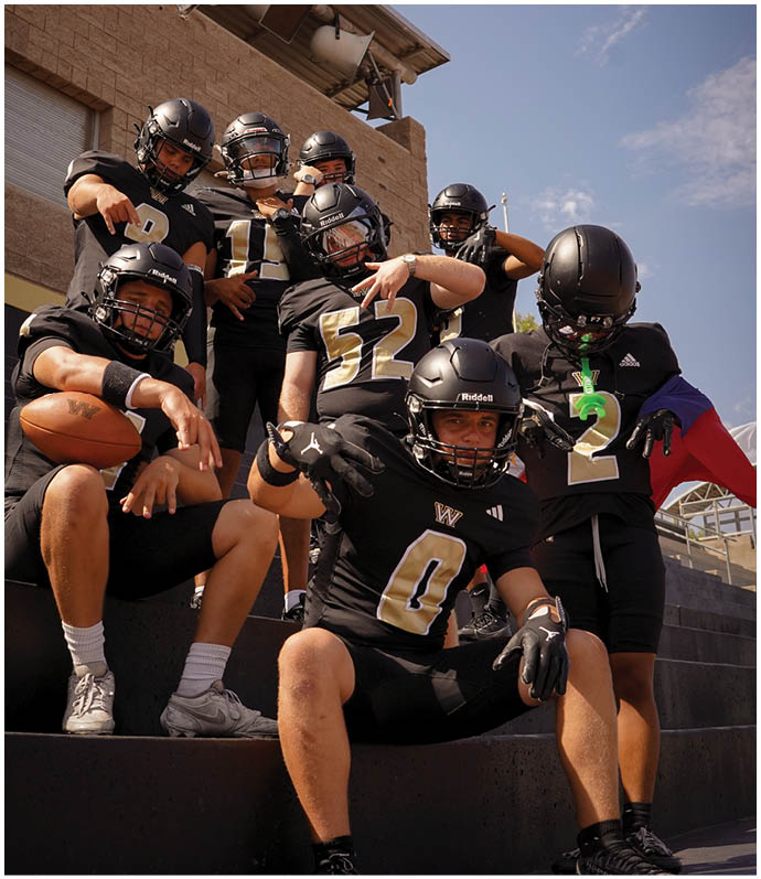 A group of seven people wearing black football uniforms and helmets pose together on stadium steps with one holding a football. Description generated by AI
