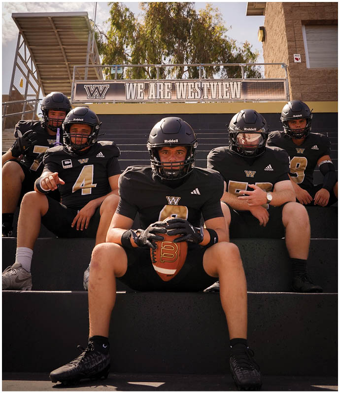 Five people wearing black football uniforms and helmets sit on stadium steps under a sign that reads “We Are Westview" with trees and a clear sky in the background. Description generated by AI