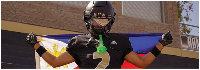 A person wearing a black football uniform and helmet stands with arms raised in front of a flag with red, blue, white, and yellow colors and a building wall. Description generated by AI