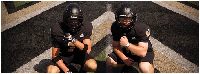 Two people wearing black football uniforms and helmets are crouching on a field with a black and white patterned surface. Description generated by AI