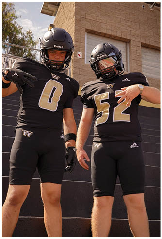 Two people wearing black football helmets and uniforms stand near a stone building with an AED sign visible on the wall. Description generated by AI