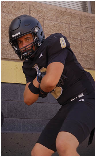 Two people wearing black football helmets and uniforms are positioned in front of a beige brick wall with two closed windows. Description generated by AI