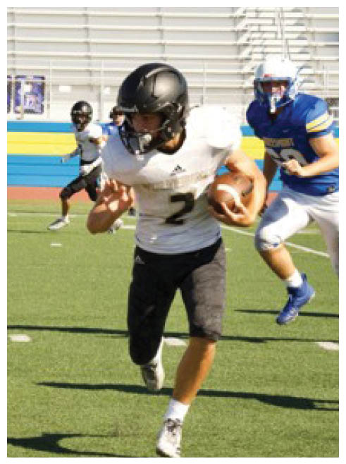 A person in a white jersey and black helmet is running with a football while people in blue jerseys attempt to block or tackle on a green field. Description generated by AI