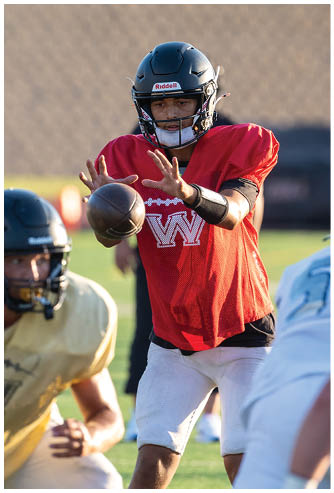 A person wearing a red sports jersey and white pants is reaching out to catch a football while two other players in helmets and sports gear are nearby on a grassy field. Description generated by AI