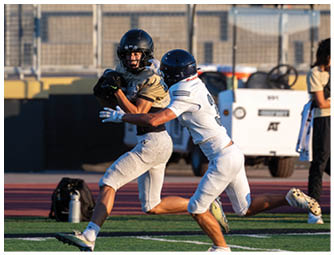 Two people wearing helmets and sports uniforms are playing football on a field, with one person running while holding the ball and the other reaching out to tackle. Description generated by AI