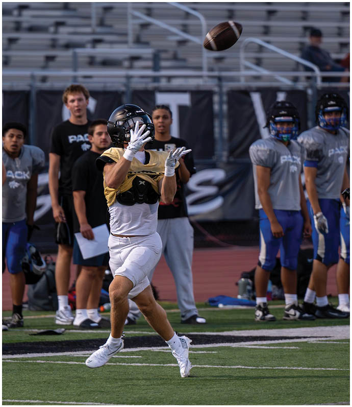 Two people in sports uniforms are actively running on a football field while others stand on the sidelines watching. Description generated by AI