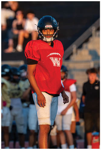 A person wearing a red football jersey and white pants stands on a field with other players and a coach blurred in the background. Description generated by AI