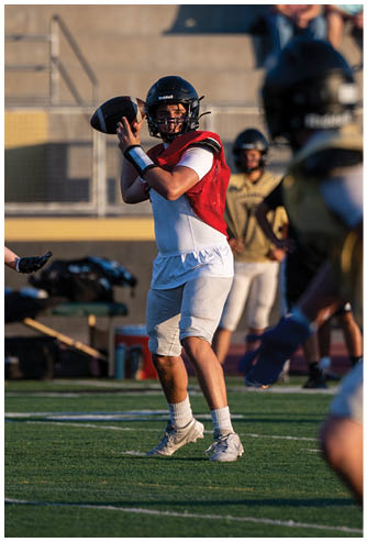 An adult wearing a red practice jersey and helmet is preparing to throw a football on a field while other adults in gold jerseys and helmets are positioned around them during a practice session with spectators in the background. Description generated by AI