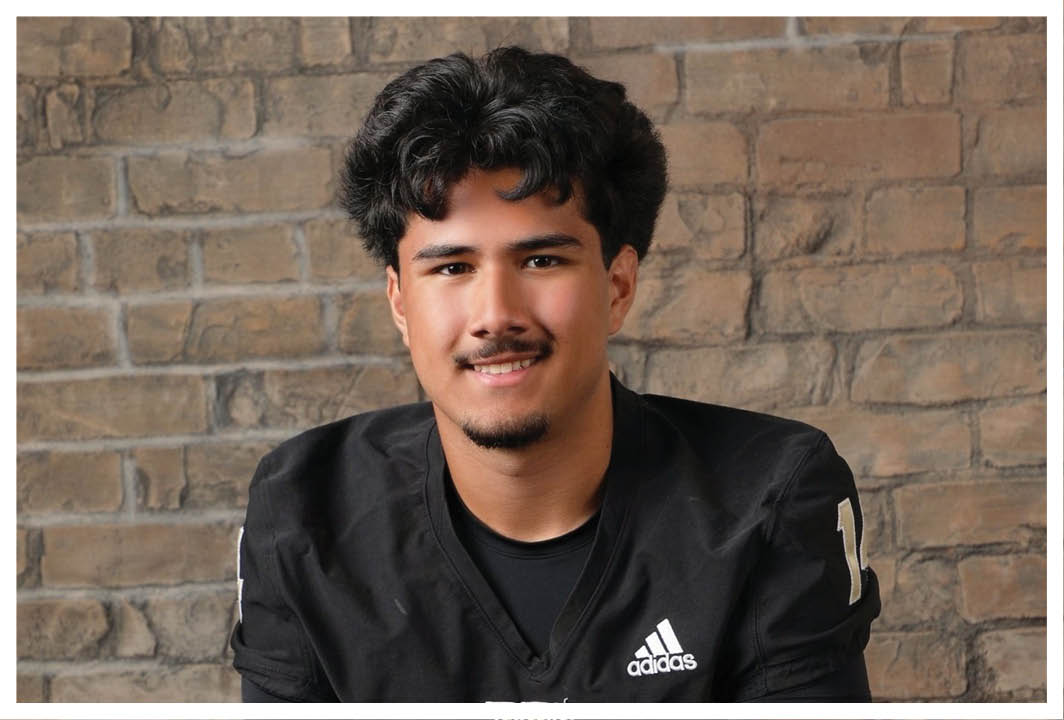 A person with curly hair wearing a black sports jersey with a “W" logo is smiling in front of a brick wall. Description generated by AI